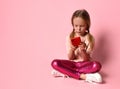 Little girl in t-shirt, leggings and sneakers. Smiling, using her red smartphone, sitting on floor with crossed legs against pink Royalty Free Stock Photo