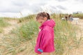 A little girl with a tail dressed in a pink raincoat stands on a sandy beach overgrown with grass. Bad weather at sea Royalty Free Stock Photo