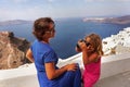 a little girl takes pictures of her mother against the beautiful sea view of Santorini