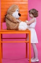 A little girl is talking to a large toy bear that is sitting on a huge chair