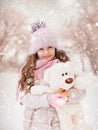 Little girl with a teddy bear stands on the street under the flying snow.