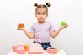 Little girl with toy hamburger and apple in hand is playing in store, toy cash register, vegetables, basket, scales, choice