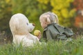 Little girl with two tails plays with teddy bear on grass near autumn forest. Fair-haired child in nature with soft toy Royalty Free Stock Photo