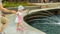 A little girl wants to swim in the fountain. Baby goes to the fountain, pulls his hand to the water. European girl in a hat walk