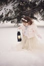 Little girl wearing a beige coat and long dress, looking at lamp with candle standing among the branches, covered with snow in the