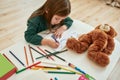 A little girl wearing green sweater sitting alone at the table with a teddy bear drawing with pencils in a large bright Royalty Free Stock Photo