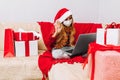 Little girl wearing medical mask  santa claus hat while talking to online friend on laptop while sitting on sofa at home for Royalty Free Stock Photo