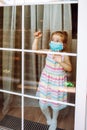 Little Girl Wearing A Medical Mask In Quarantine Staying Behind Big Window And Waiting For Her Parents From Work. Coronavirus