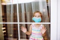 Little Girl Wearing A Medical Mask In Quarantine Staying Behind Big Window With Wide Opened Eyes. Coronavirus Quarantine Concept