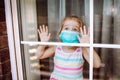 Little Girl Wearing A Medical Mask In Quarantine Staying Behind Big Window With Wide Opened Eyes. Coronavirus Quarantine Concept.