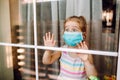 Little girl wearing a medical mask staying behind big window with wide opened eyes. Coronavirus quarantine concept. Outbreak Royalty Free Stock Photo