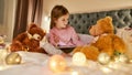 Little girl wearing pyjamas sitting alone on a huge bed barefoot with her tablet and speaking to her fluffy teddybear Royalty Free Stock Photo