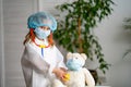 Little girl in white coat cap and mask, using stethoscope, listens to Teddy bear