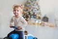 Little girl in a white sweater rides a toy vintage blue car. Christmas tree in the background, gifts, lights Royalty Free Stock Photo