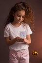 A little girl in a white t-shirt with curly long hair, holding a ball of Christmas tree decoration, looks away on a dark purple Royalty Free Stock Photo