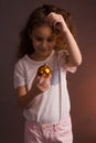 A little girl in a white t-shirt with curly long hair, holding a ball of Christmas tree decoration, looks away on a dark purple Royalty Free Stock Photo