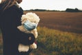 little girl with white teddy bear wearing a face mask observes her sorrounding wistfully outdoors longing for social contacts