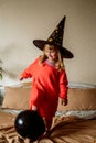 Little girl in a witch hat with an orange and black ball . Active games at home. Laughs emotionally and jumps on the bed.