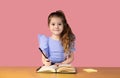 A little girl writes in a notebook and holds a pen in her right hand, she is surprised in the school desk on a pink Royalty Free Stock Photo