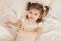 Little girl 3 years old lies cheerful with smartphone in her hands on white bed, lifestyle