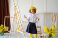 little girl in yellow beret and tights stands near the easel while holds color pallete and brush
