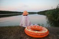 A little girl in a yellow hat, covered with a towel, sits on the shore of the lake, next to an inflatable circle, summer evening- Royalty Free Stock Photo