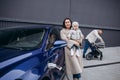 Little girl and young woman standing by trunk while charging electric car at station.