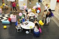 Little girls and adults sitting at tables painting at children lounge zone of a shopping mall.