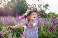 A little happy blonde girl in a sage wreath and a colorful dress in a summer blooming field