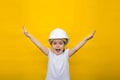 Little happy girl in a construction white helmet hands up on a yellow background