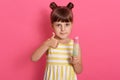 Little happy kid girl holding ice cream and showing thumb up while looking directly at camera, wearing summer dress, has two knots Royalty Free Stock Photo