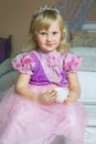 Little happy princess girl in pink dress and crown in her royal room sitting on chair and holding cup of drink. Royalty Free Stock Photo