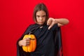 Little hispanic boy wearing a cape and holding halloween pumpkin with angry face, negative sign showing dislike with thumbs down, Royalty Free Stock Photo