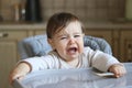 Little hungry baby crying sitting in the high feeding chair with spoon in his hand, Royalty Free Stock Photo