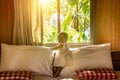 A little infant baby girl in a white dress is standing on a bed against the background of the window behind which the jungle. A Royalty Free Stock Photo