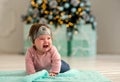 Little infant girl lying or crawling on floor in room with decorated Christmas tree and crying. Winter holidays with little kids