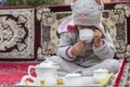 Little Iranian girl in a hat in the form of a kitty is drinking tea on a sofa in a traditional Persian coffee shop