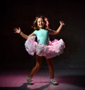 Little kid in blue t-shirt and poofy skirt. Smiling, closed eyes, jumping up against black background, pink backlight. Close up