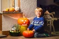 Little kid boy sitting with traditional jack-o-lanterns pumpkins for halloween by the decorated scary door, outdoors