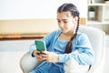 Little kid cute girl sitting on chair, having fun at home, using smartphone, playing video game on mobile phone. Royalty Free Stock Photo