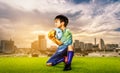 Kid goalkeeper holding the ball on a grass field with golden sky city background for success soccer dramatic concept Royalty Free Stock Photo