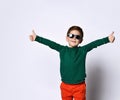 Little kid in sunglasses, green jumper and orange pants. He is smiling and showing thumbs up, posing isolated on white Royalty Free Stock Photo