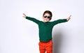 Little kid in sunglasses, green jumper and orange pants. He is smiling and showing thumbs up, posing isolated on white Royalty Free Stock Photo