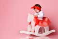 Little kid in white bodysuit, red headband and poofy skirt. She is riding a rocking horse against pink studio background. Close up
