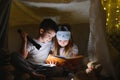 Little kids involving in reading amazing book. They lying in nice toy tent in playroom. Boy holding flashlight in hand.