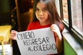 Little latin girl standing near the window and holding a sheet of paper that says in spanish `I want to go back to school`. Royalty Free Stock Photo