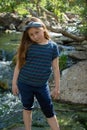 Little latina girl smiling coyly and looking down while standing in front of a stream and woods in the shade