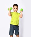 Little male in yellow t-shirt and denim shorts. He smiling, lifting green dumbbell, posing isolated on white studio background Royalty Free Stock Photo