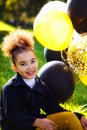 A little mixed-race girl is playing outdoors in an autumn park. He holds multicolored gold, black, and silver balloons in his