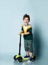 Little model in colorful t-shirt, gray jeans and sneakers. He is screaming standing on kick scooter against blue background